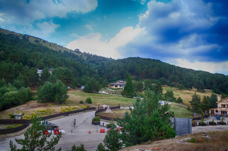 A pine forested mountain pass with a curving road, green meadow and mountain buildings at its end