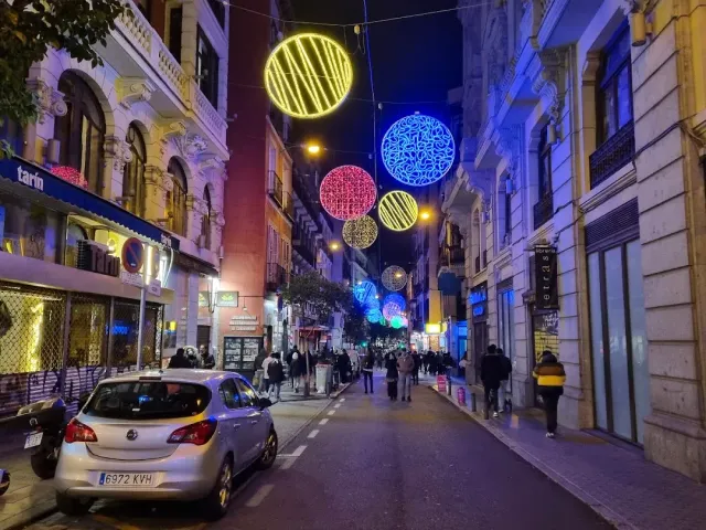 An evening street with round hanging light decorations, lit shops and passersby