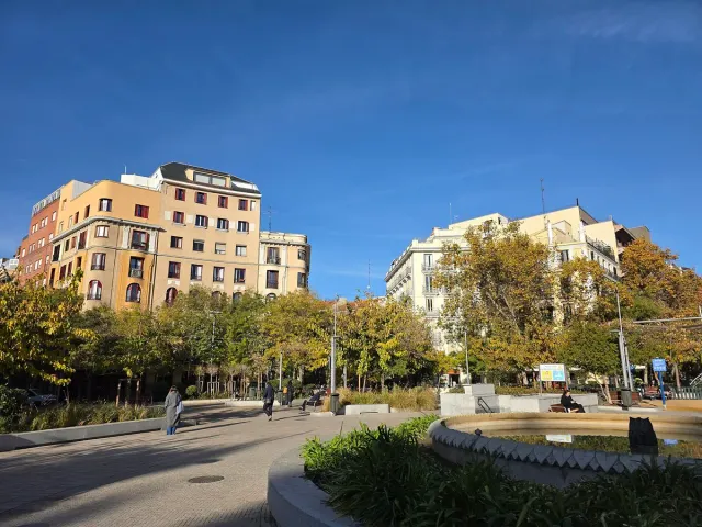 A round square with a stone fountain, benches and autumn trees in front of pale apartment blocks