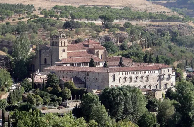 A stone monastery with a tall bell tower in a green valley below a built up ridge