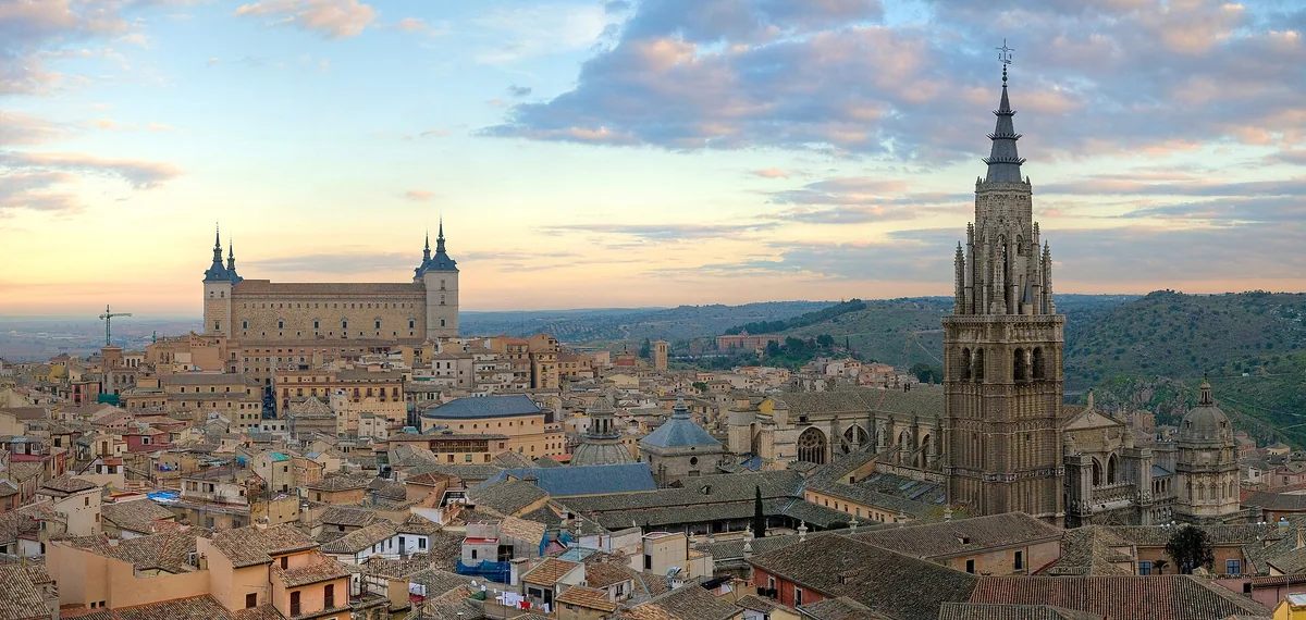 A view over the tiled roofs of an old hilltop city, with a gothic cathedral tower and a square fortress in evening light