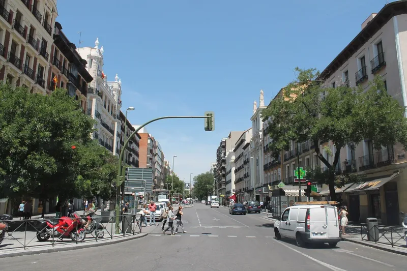 A wide city street in daylight with balconied buildings on both sides, shops and pedestrians crossing