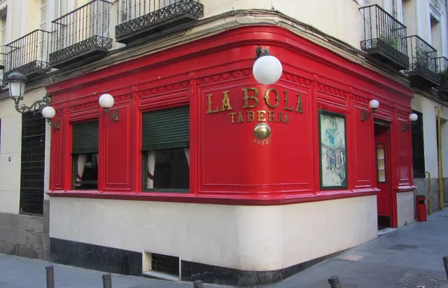 A bright red corner tavern front with a gilded sign and round lamps