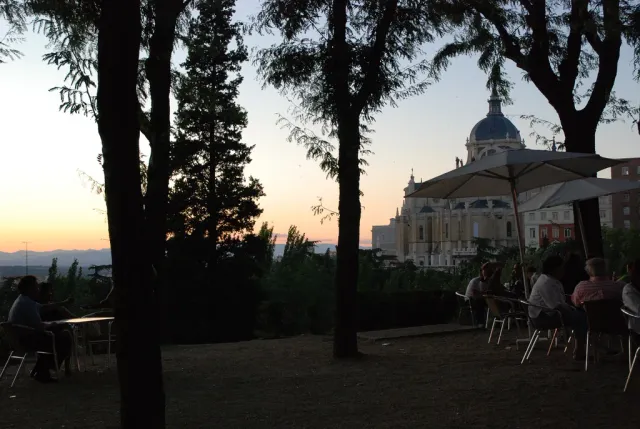 A terrace with tables among trees at sunset, with a cathedral dome visible beyond the rooftops