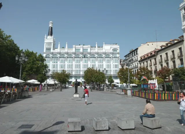 A wide paved square with cafe terraces and an ornate white building with a spire at its far end