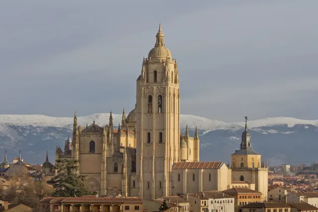 A pale stone Gothic cathedral with a tall bell tower against a snow covered mountain ridge