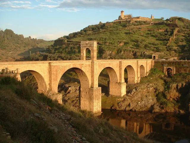 An ancient stone bridge with wide arches over a river, with a fortress on the hill behind