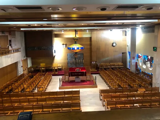 A hall with rows of wooden benches, a raised platform with a red carpet and a hanging stained glass lamp