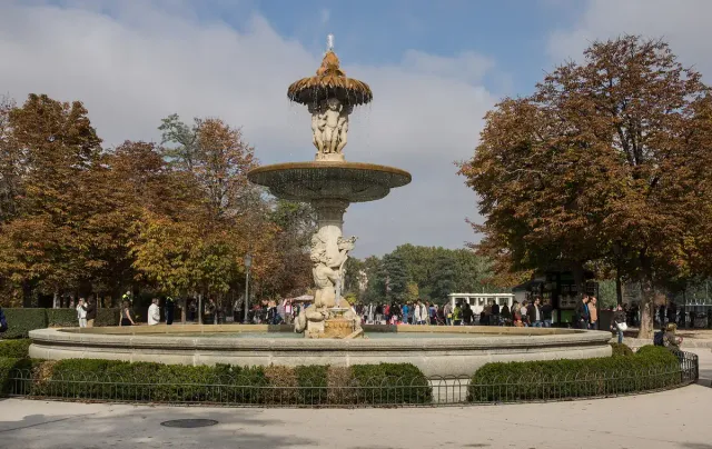 A tiered stone fountain with an upper basin and statues at its base in a garden with trees