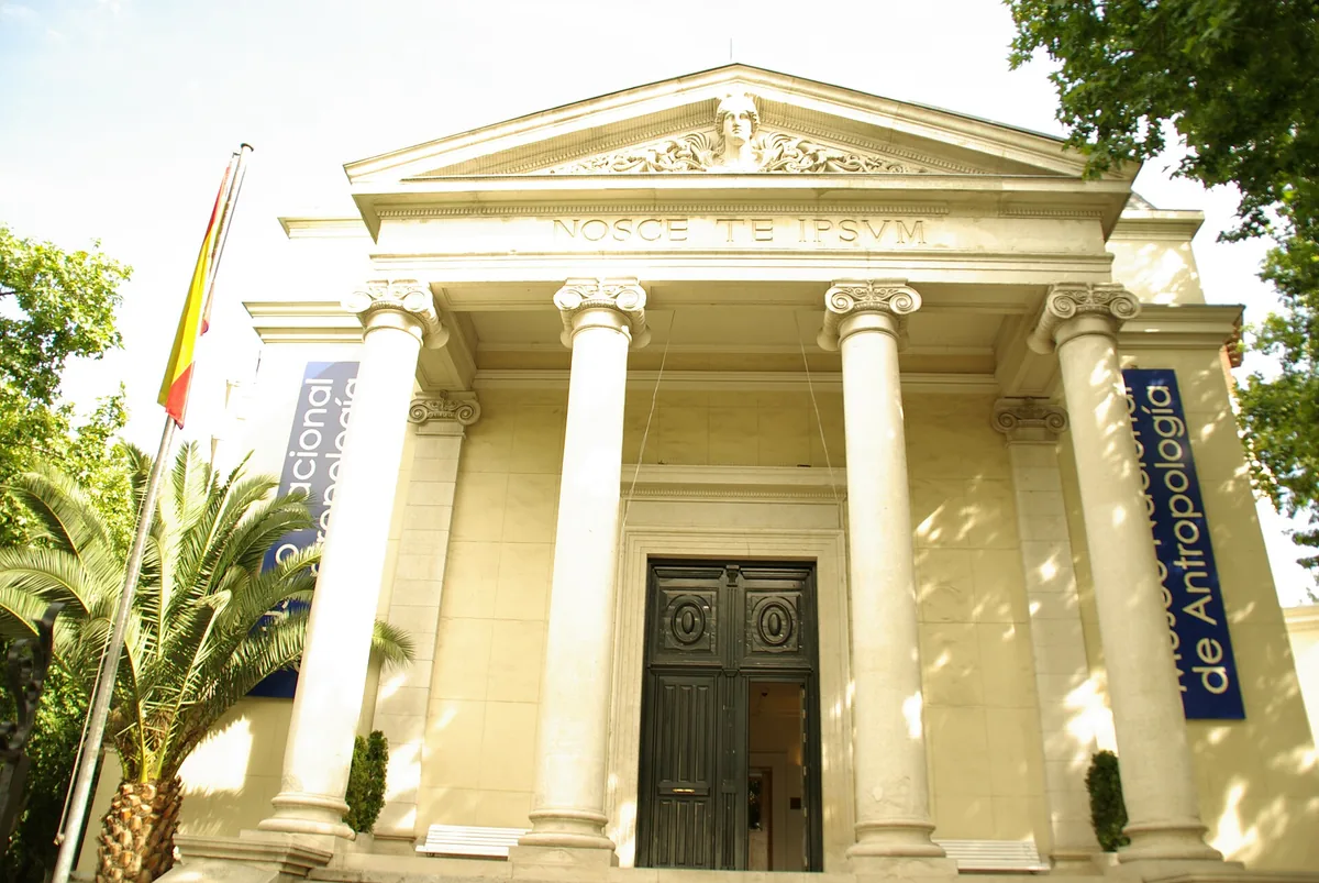 A neoclassical entrance portico with columns and a carved pediment, a palm and blue banners beside it