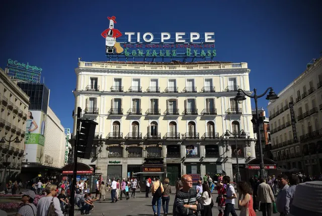 A large bottle shaped advertising sign with a red hat on a rooftop above a crowded square