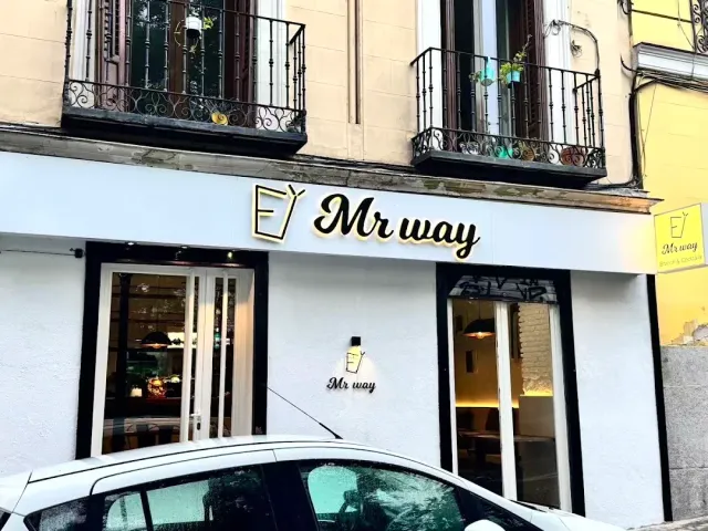 A white frontage with a gilded sign and iron balconies above it, a car parked in front