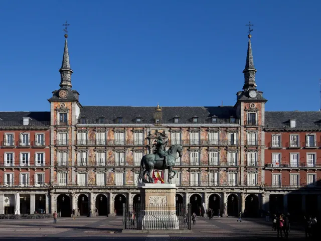 A symmetrical red facade with two spires and painted murals, and a bronze equestrian statue in the middle of the square
