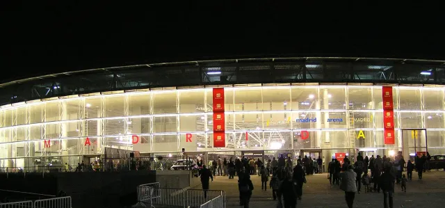 A curved events hall facade lit up at night with a crowd entering through the gates