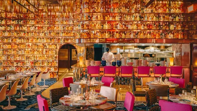 A restaurant room with walls covered in illuminated bottles, an open kitchen and pink chairs