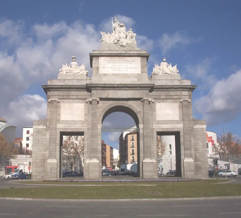 A neoclassical stone triumphal gate with sculpture groups on its roof, standing on a traffic island against a blue sky