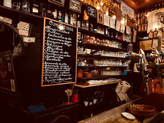 A dark tavern interior with a written chalkboard, shelves of glasses and bottles and drink taps