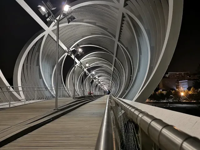 A pedestrian bridge inside a lit metal spiral at night, with a wooden floor and a railing