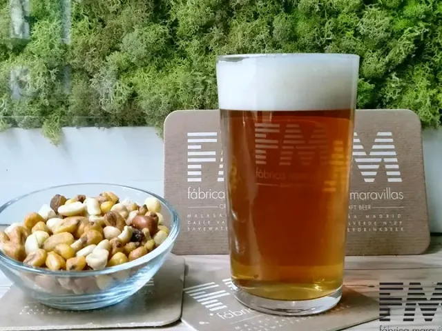 A glass of pale beer with foam beside a small bowl of nuts against a green wall
