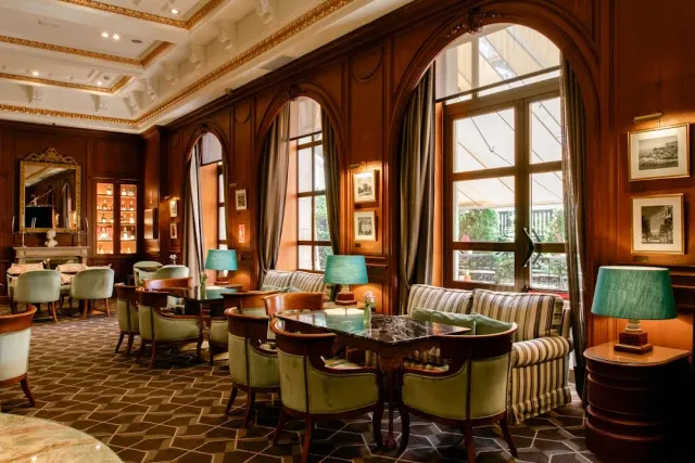 A wood panelled lounge with green armchairs, table lamps and arched windows