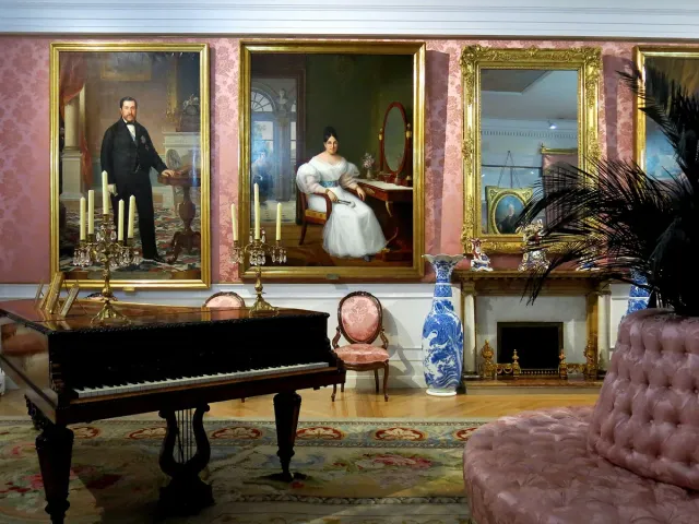 A nineteenth century drawing room with a grand piano, a pink sofa and large portraits on the walls