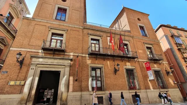 A brick palace facade with iron balconies and flags, and passers by on the pavement
