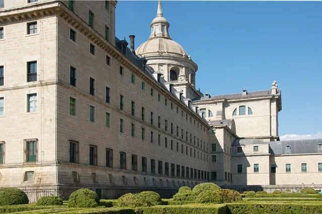 A long stone monastery wing with a tall dome above it and clipped hedges in front