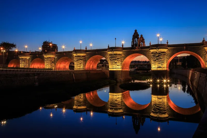 A many arched stone bridge lit orange above still water, with its arches reflected and a deep blue dusk sky