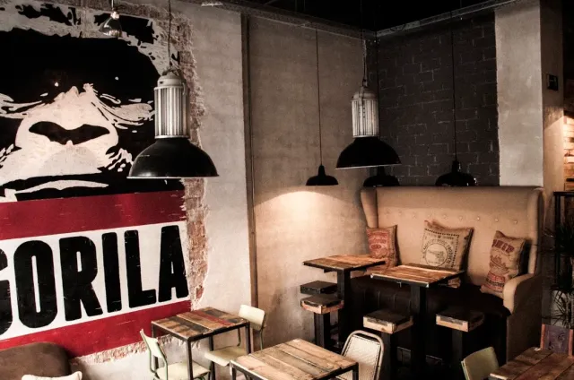 A black and white wall painting of an ape face above wooden tables and metal lamps