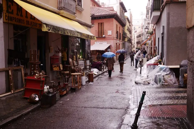 An open air market street on a rainy day with stalls, goods on tables and shoppers under umbrellas