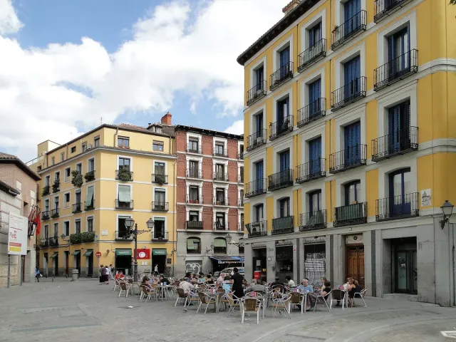 An open square ringed by yellow and blue balconied houses, with parasol terraces in the middle