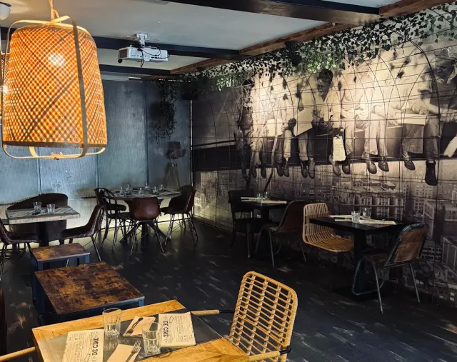 A dark dining room with a large black and white photographic mural, a wicker lamp and wooden tables