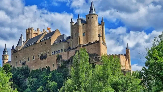 A stone castle with pointed spires on top of a cliff, above a green grove of trees