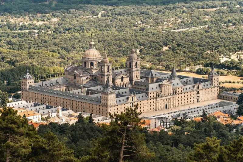 A vast monastery building with a dome and towers, ringed by forest with red tiled village roofs below