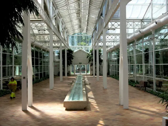 The interior of a glass and iron greenhouse with a long water channel and white columns on either side