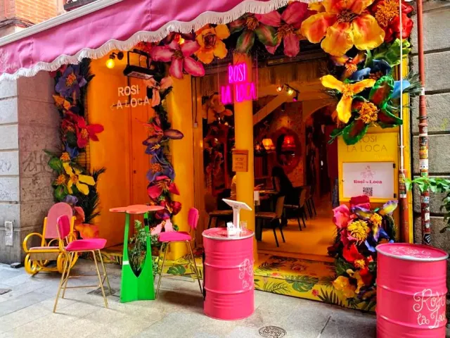 A colourful facade with giant fabric flowers, a pink awning and pink barrels at the entrance