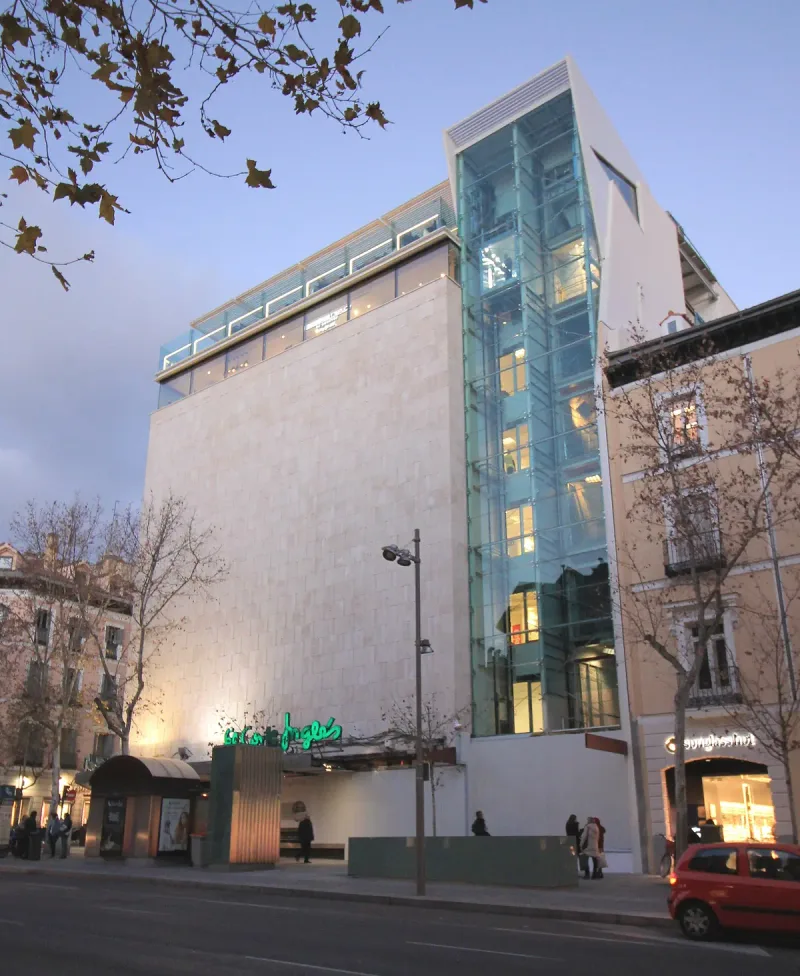 A pale stone department store building with a glass lift tower and a green El Corte Ingles neon sign at dusk