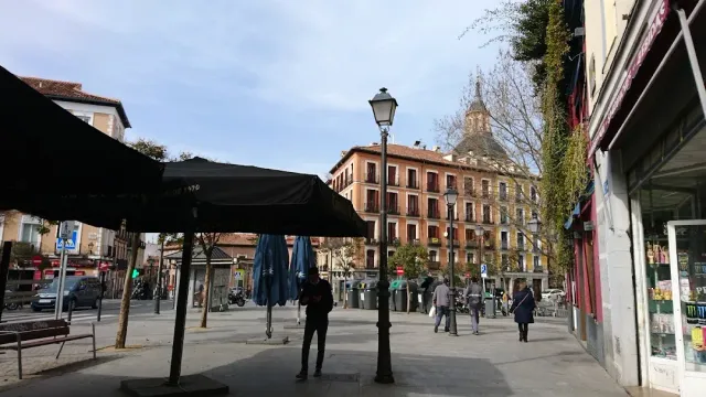 A paved square with folded terrace umbrellas, a street lamp and passers by in front of residential buildings