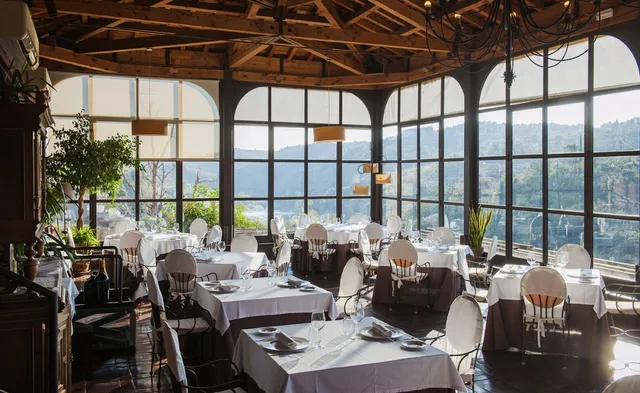 A wide dining hall with white tablecloths, a wooden beamed ceiling and arched windows onto green hills