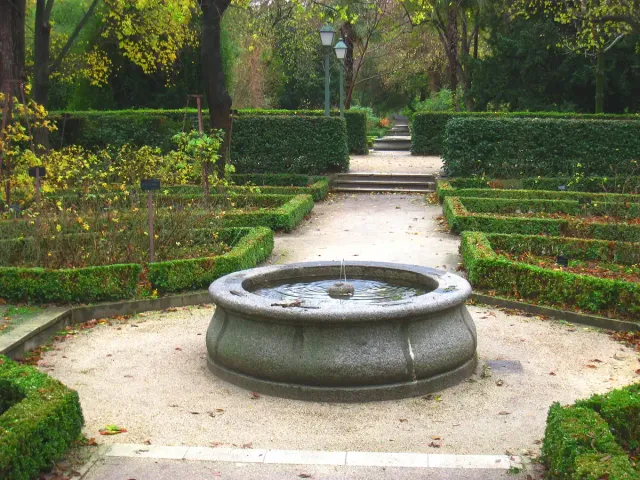 Beds edged with clipped hedges around a round stone basin, on gravel paths among trees