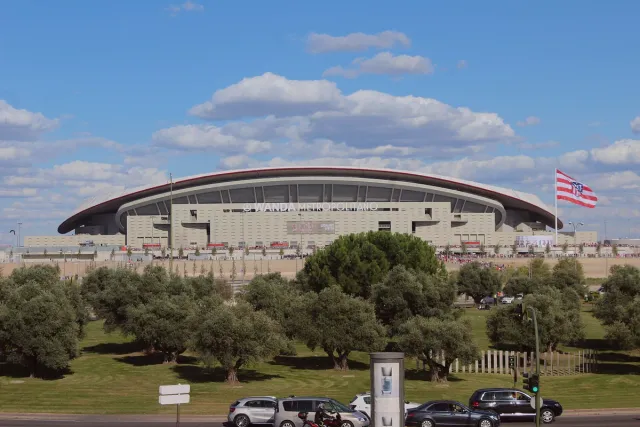A large stadium with an undulating white roof, surrounded by an open forecourt and flags