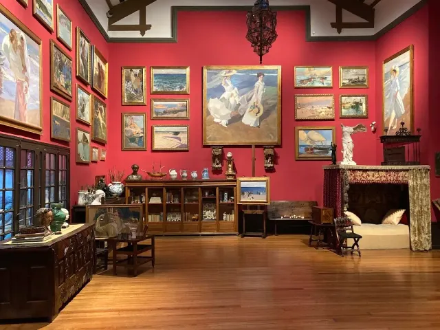 A room with red walls covered in framed paintings, a wooden floor and a display cabinet