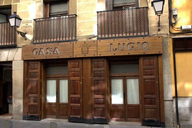 A dark wooden restaurant front with a gilded lettered sign, lamps and lace curtains in the windows