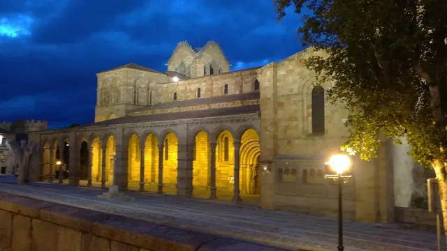 A stone basilica with an arcaded porch lit in orange against a deep blue evening sky