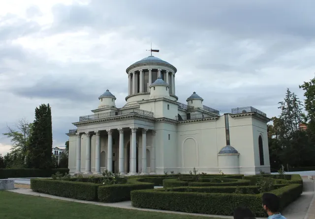 A small neoclassical building with a columned porch and a dome on a hill surrounded by a tidy garden