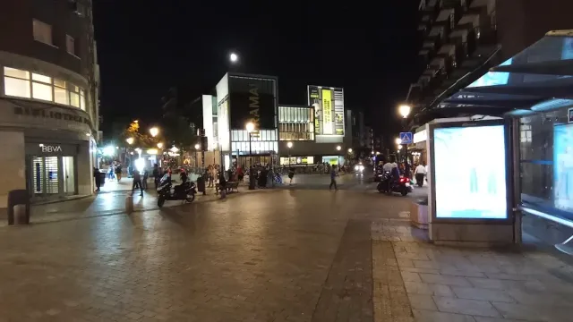 A paved plaza at night with passers by, parked scooters and illuminated signs