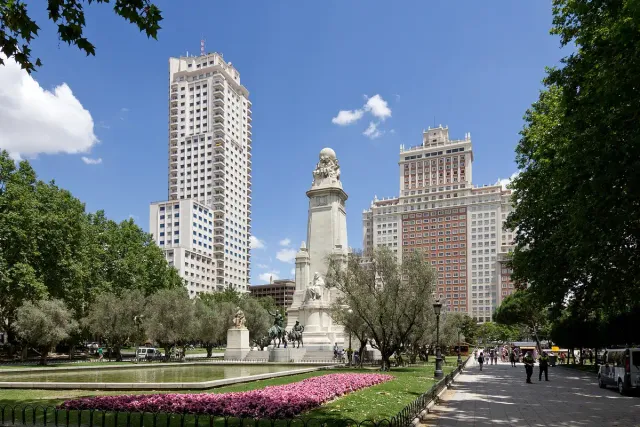 A square with a long pool and a stone monument, a pink flower bed and tall towers behind