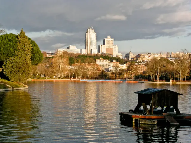 A calm lake with a small wooden jetty and a city skyline beyond the trees