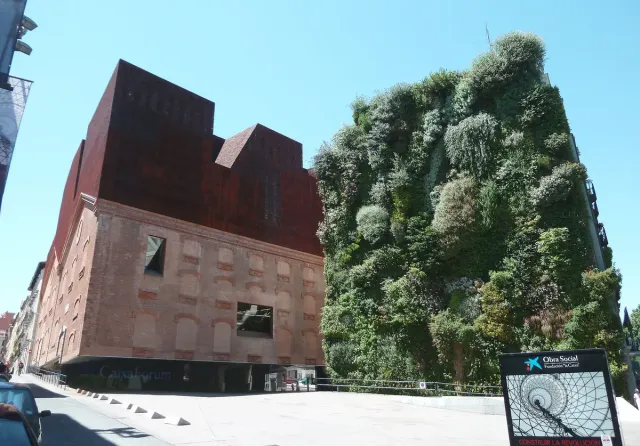 A rust clad building floating above a stone base, beside a wall covered in green planting