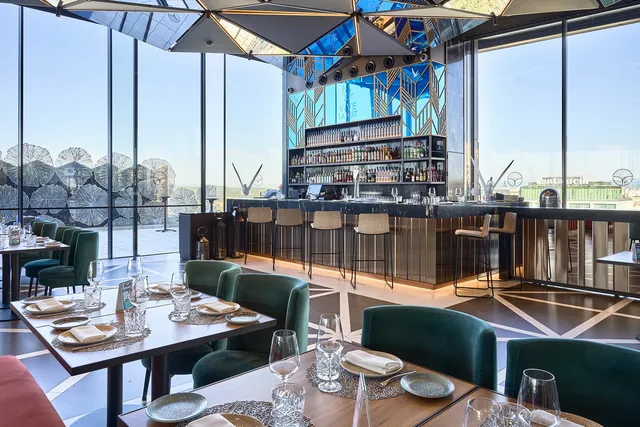 A bright glass-walled restaurant with green armchairs and a long bar facing the city view